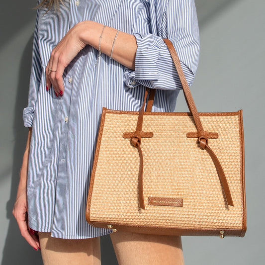 Model holds a rectangular straw effect bag with brown leather trim and handles.