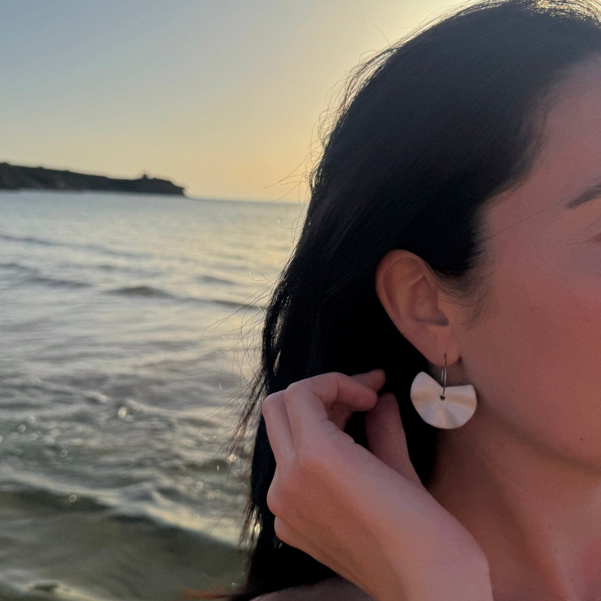 Close-up of a woman wearing Frolla white ceramic dangle earrings by the sea at sunset