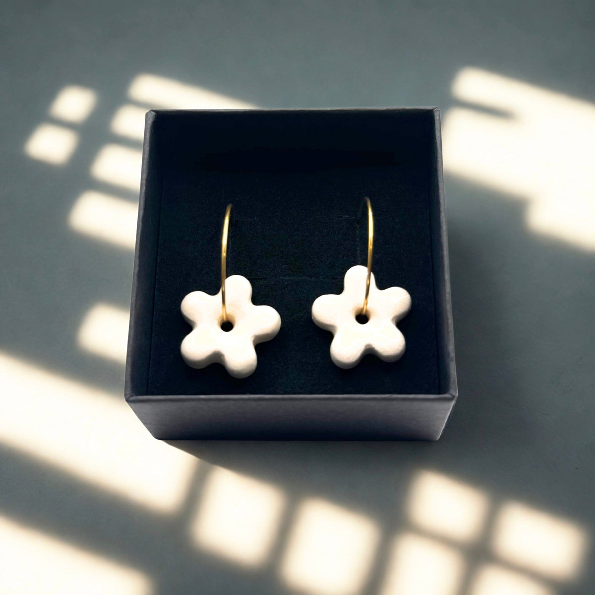Five-petal white ceramic flower earrings on gold hoops in a dark jewelry box
