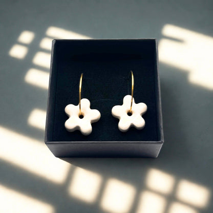 Five-petal white ceramic flower earrings on gold hoops in a dark jewelry box