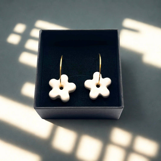 Five-petal white ceramic flower earrings on gold hoops in a dark jewelry box