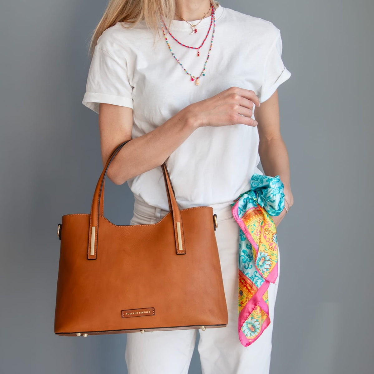 Person holding a marrone leather tote hebag con a colorful scarf draped over their arm against a grigio background
