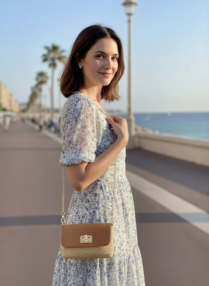 Woman in a blue floral dress on a seaside promenade wearing a raffia and leather shoulder bag with a gold chain strap.