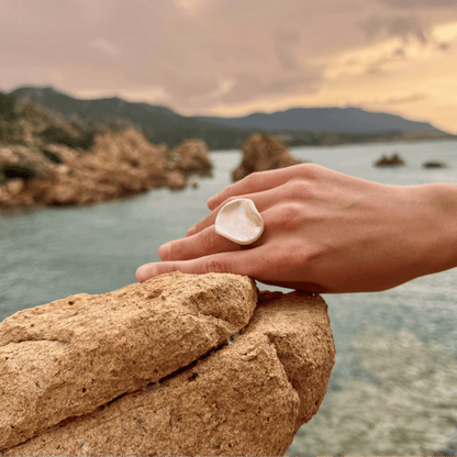 Pearl white ceramic ring on a hand resting on a rocky ledge by the sea at sunset.