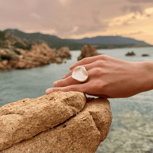 Pearl white ceramic ring on a hand resting on a rocky ledge by the sea at sunset.