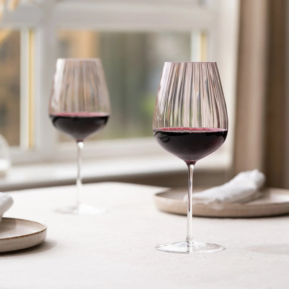 Zwei staubrosa gerippte Weingläser mit Rotwein auf einem Tisch