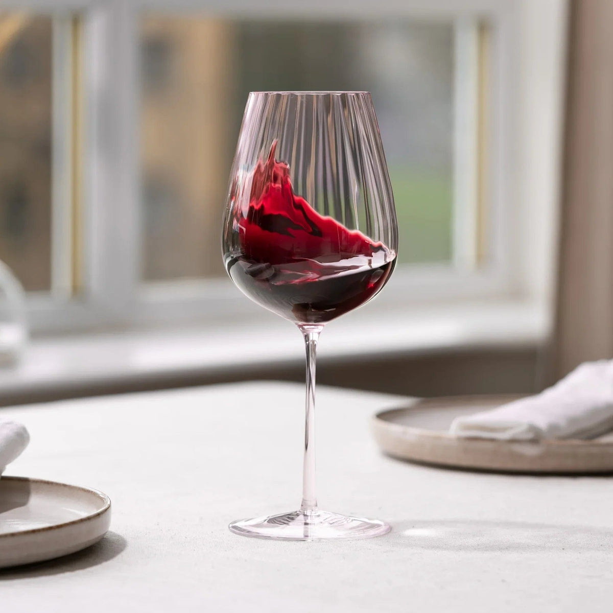 Staubrosa geripptes Weinglas mit Rotwein auf einem Esstisch nahe einem Fenster