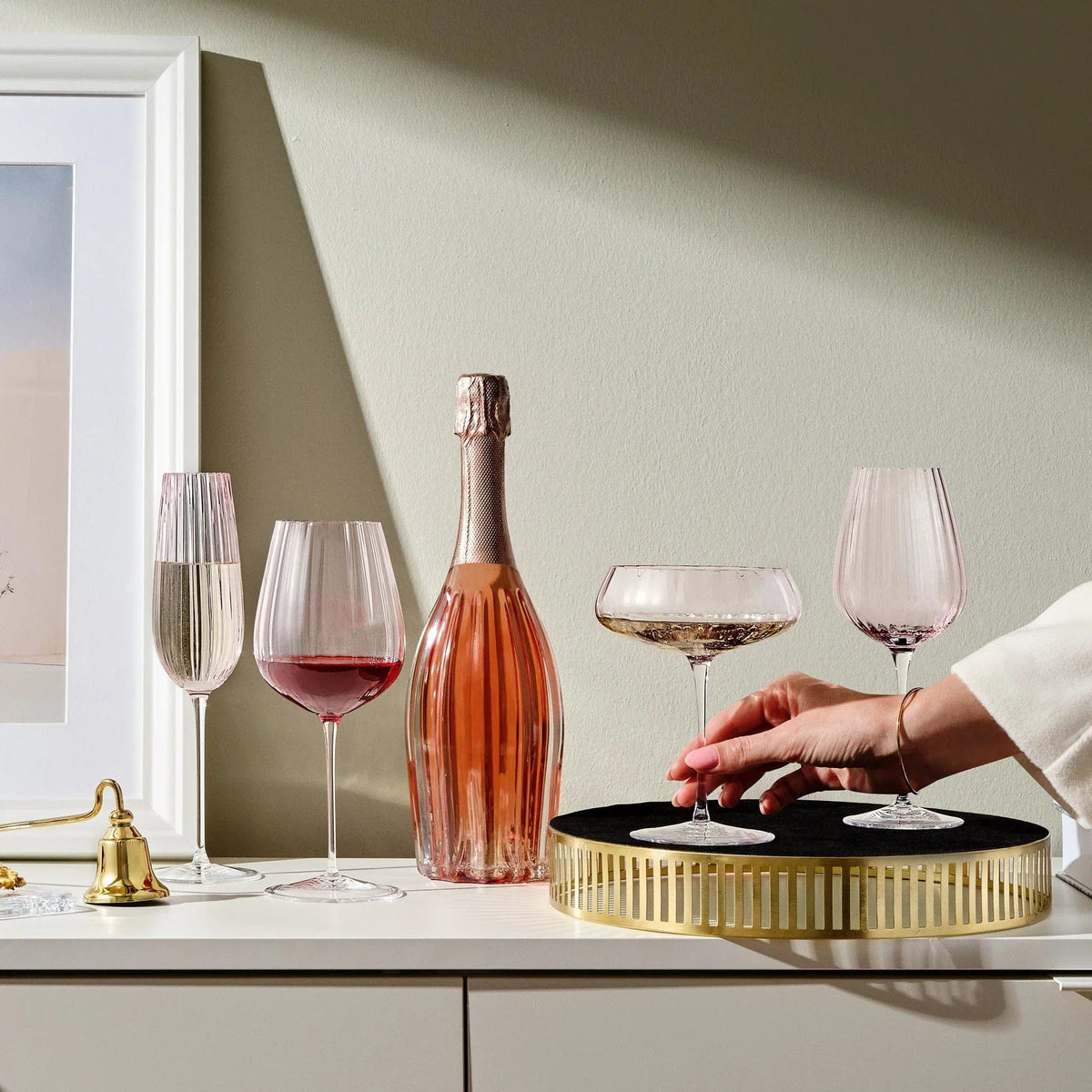 Two dusty rose white wine glasses beside a champagne bottle on a white sideboard