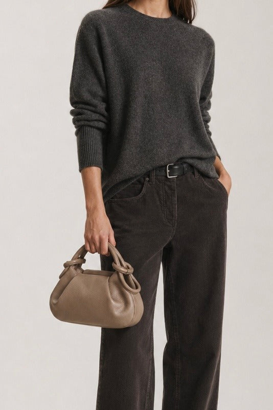 A model wearing a dark charcoal gray knit sweater and dark brown wide-leg corduroy trousers, holding a small taupe leather handbag against a light gray studio background.