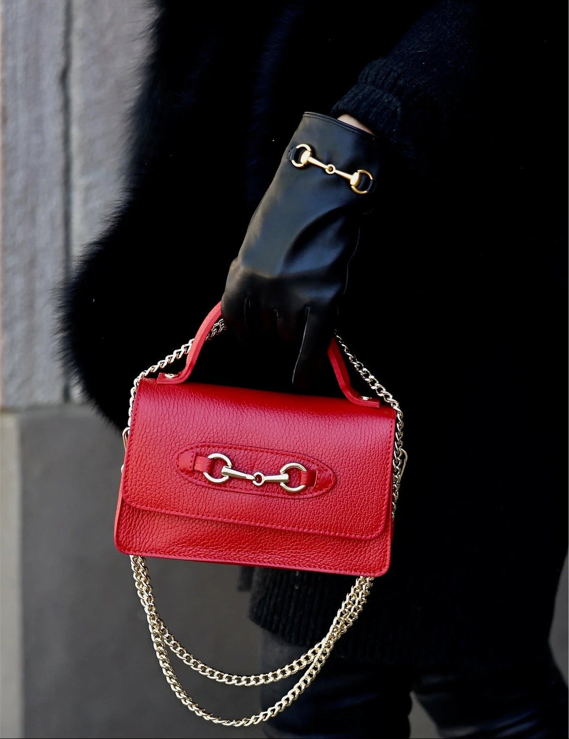 Red handbag with gold hardware held by a gloved hand against a blurred background