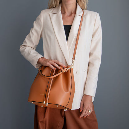 A woman wearing a cream blazer and brown pants carries a tan leather bucket bag with a matching shoulder strap and gold-tone hardware against a dark gray studio background.