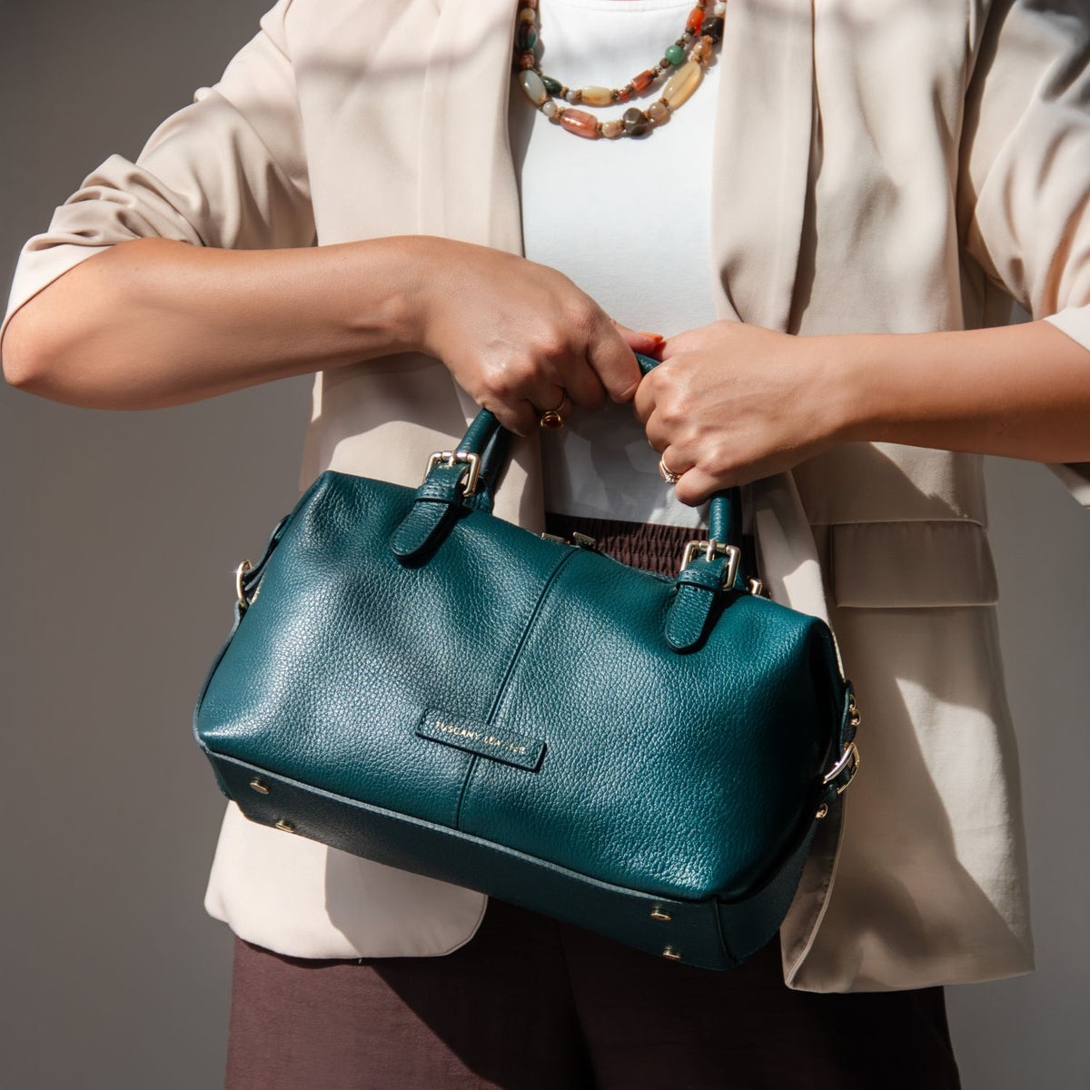 A woman in a beige blazer and white top holds a teal pebbled leather handbag with gold-tone hardware. The brand name 'TUSCANY LEATHER' is visible on a small leather label on the front of the bag.
