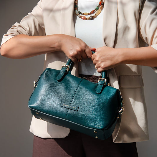 A woman in a beige blazer and white top holds a teal pebbled leather handbag with gold-tone hardware. The brand name 'TUSCANY LEATHER' is visible on a small leather label on the front of the bag.