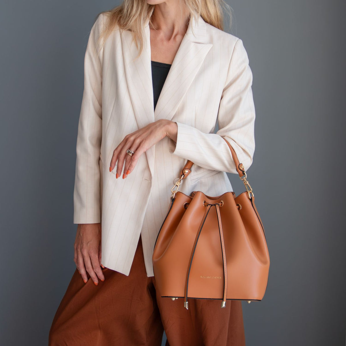 A model wearing a cream blazer and brown trousers carries a tan leather drawstring bucket bag with gold-tone hardware against a dark gray studio background.