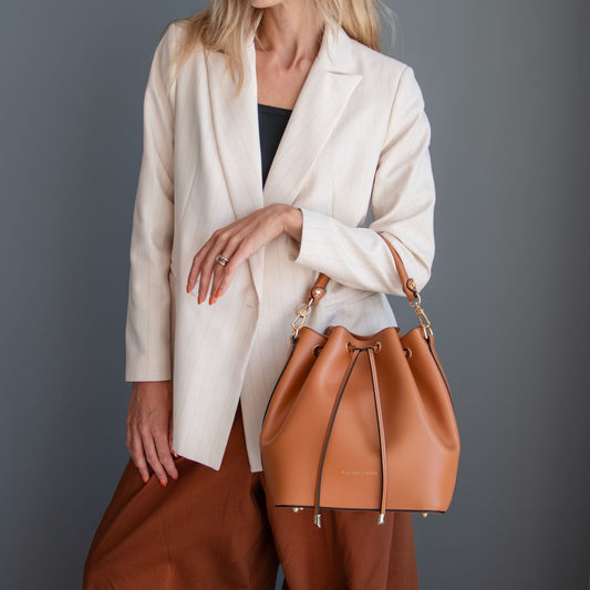 A modelo con a cream blazer and brown trousers carries a cuero leather drawstring bucket bag with herrajes dorados against a dark gray studio background.