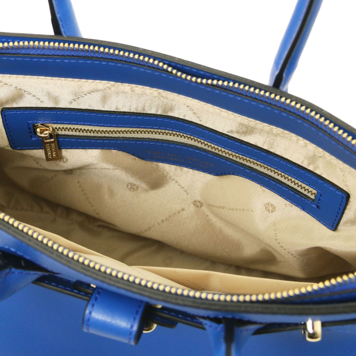 Abierto top view of blue leather handbag revealing beige forro interior with bolsillo con cremallera and gold-tone zipper pull.