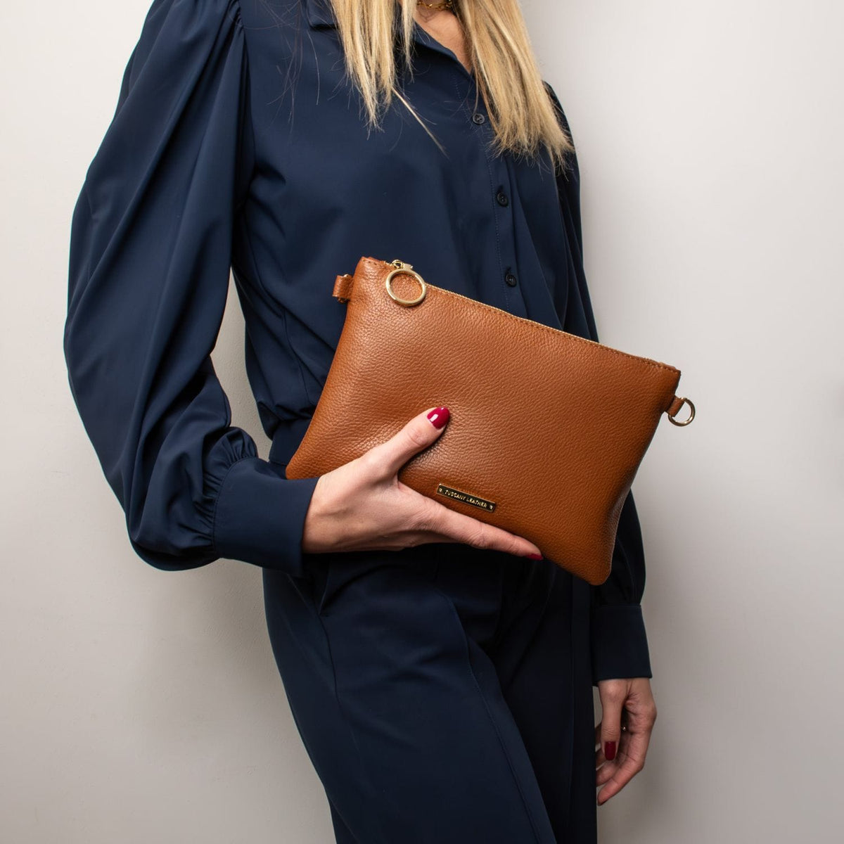 Woman in a navy blouse holding a tan soft Italian leather pouch clutch with gold ring hardware and a small plaque.