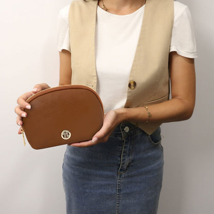Rounded cuero brown piel suave toiletry case with cremallera dorada sostenido por una persona in a white top.