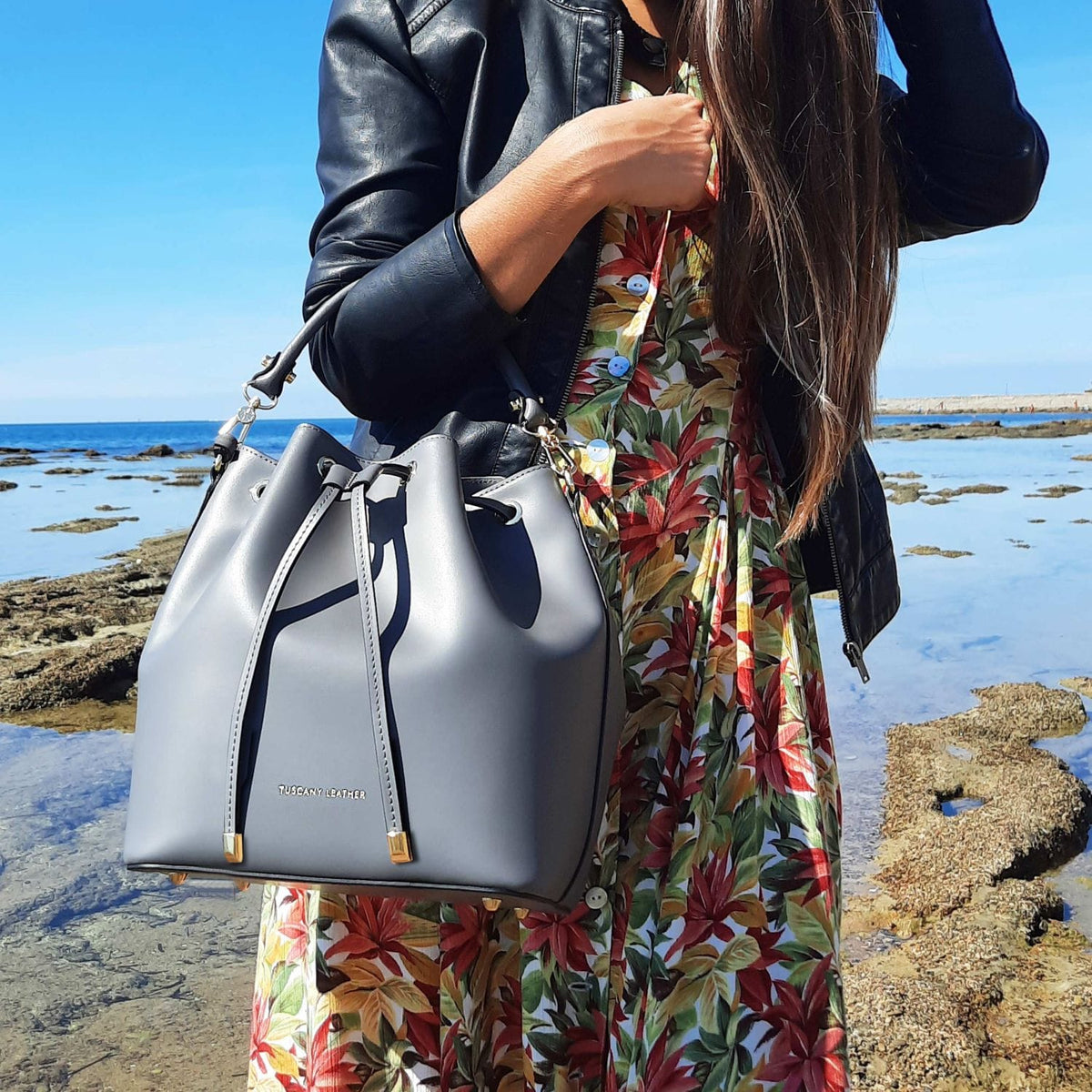 Vittoria Italian leather bucket bag in blue gray carried by a woman in a floral dress beside the sea.